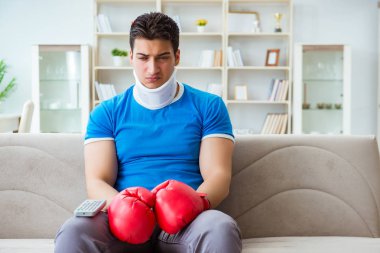Boynu yaralanmış bir adam evde boks seyrediyor.