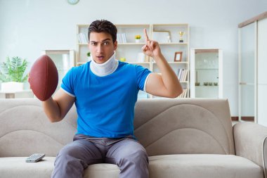 Boynu yaralanmış bir adam evde Amerikan futbolu seyrediyor.