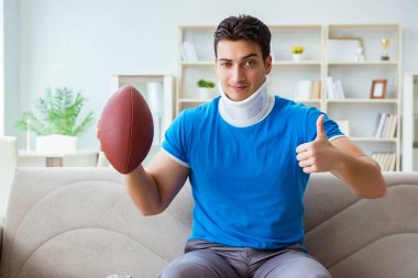 Boynu yaralanmış bir adam evde Amerikan futbolu seyrediyor.