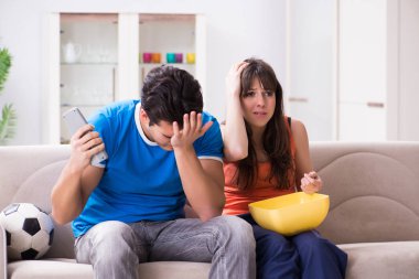Genç adam evde karısıyla futbol seyrediyor.