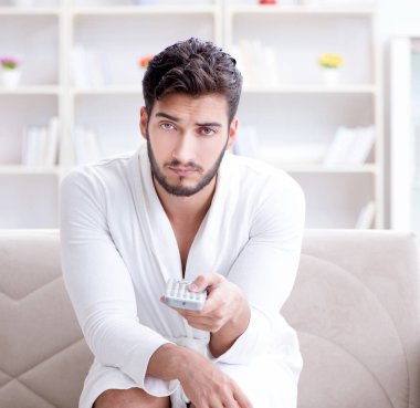 Bornozlu genç adam evde kanepede televizyon izliyor.