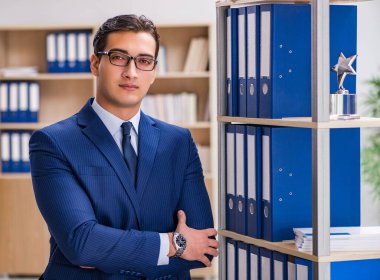 Dosyalarla rafın yanında duran genç adam.