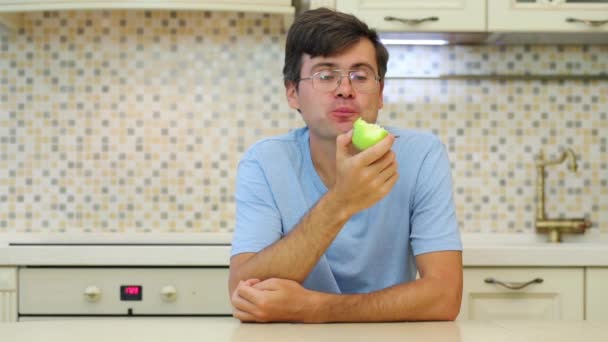 戴眼镜和蓝色 T恤的男士吃绿色苹果坐在厨房里 — 图库视频影像