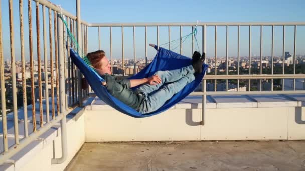 屋根の上の手すりに接続されているハンモックに横になっているジーンズの男 — ストック動画