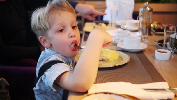 テーブルでレストランでフォークで食べるブロンドの髪を持った少年 — ストック動画