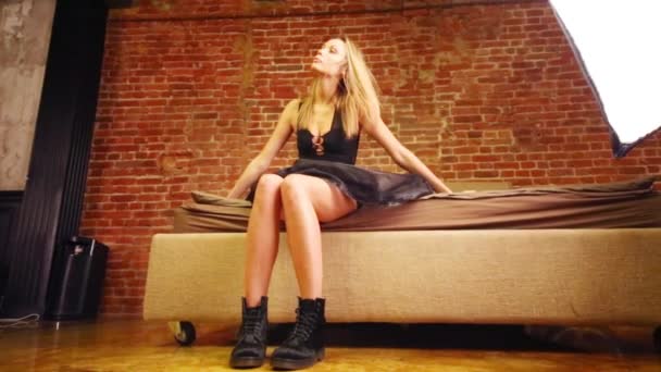穿着黑色连衣裙的漂亮金发女郎坐在工作室的床上 — 图库视频影像