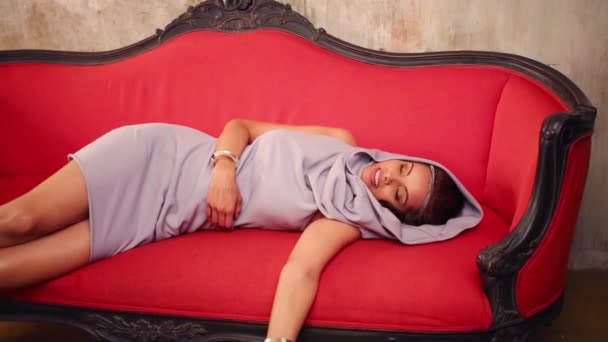 スタジオの赤いソファに位置してフード付きのドレスを着た美しい女性 — ストック動画