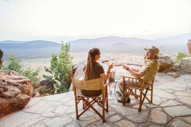 Romantic couple on safari vacation sitting outdoor at lodge enjoying stunning views over national park with glass of sundowner wine clipart