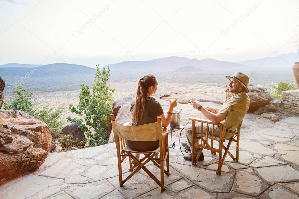 Romantic couple on safari vacation sitting outdoor at lodge enjoying stunning views over national park with glass of sundowner wine