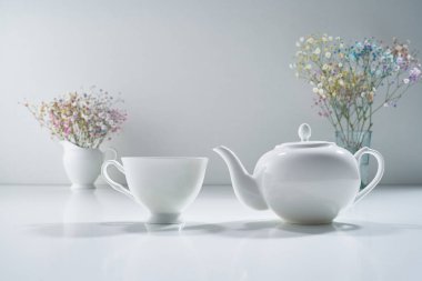 Açık gri arka planda beyaz porselen çay takımı ve narin bir buket çiçekle kompozisyon