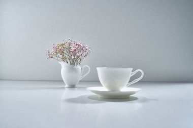 Açık gri arka planda beyaz porselen çay takımı ve narin bir buket çiçekle kompozisyon