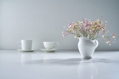 Açık gri arka planda beyaz porselen çay takımı ve narin bir buket çiçekle kompozisyon