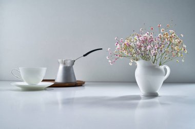 Açık gri arka planda beyaz porselen çay takımı ve narin bir buket çiçekle kompozisyon