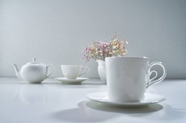Açık gri arka planda beyaz porselen çay takımı ve narin bir buket çiçekle kompozisyon
