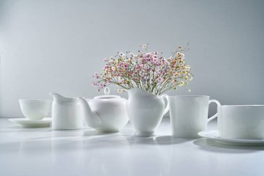 Açık gri arka planda beyaz porselen çay takımı ve narin bir buket çiçekle kompozisyon