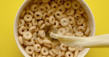 Süt, beyaz bir fincana dökülür. Ağız sulandıran mısır gevreği halkaları parlak sarı bir zemin üzerindedir.