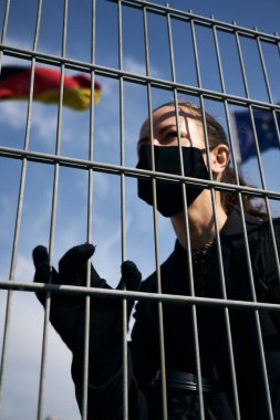 Yüzünde siyah koruyucu bir maske olan açık tenli sarışın kız blok ızgarasının arkasında duruyor. Arka planda dalgalanan bayrak.