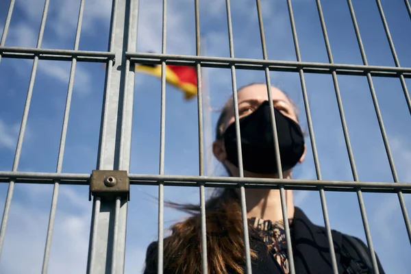 Light-skinned blonde girl in a black protective mask on her face, stands behind the blocking grate, the flag flutters in the background