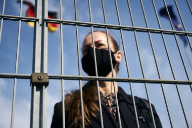 Yüzünde siyah koruyucu bir maske olan açık tenli sarışın kız blok ızgarasının arkasında duruyor. Arka planda dalgalanan bayrak.
