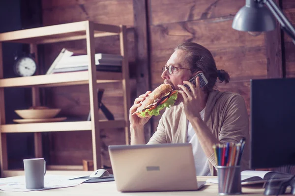 Freelancer comer sanduíche enquanto fala por telefone . — Fotografia de Stock