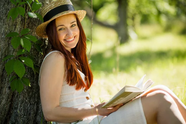 Ρομαντική κοπέλα σε ψάθινο καπέλο διαβάζοντας ένα βιβλίο που κάθεται στον κήπο. — Φωτογραφία Αρχείου