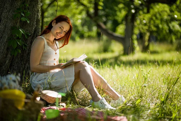 Vonzó ravasz nő ül vele vissza, közel a fa a kertben könyvet olvas. — Stock Fotó