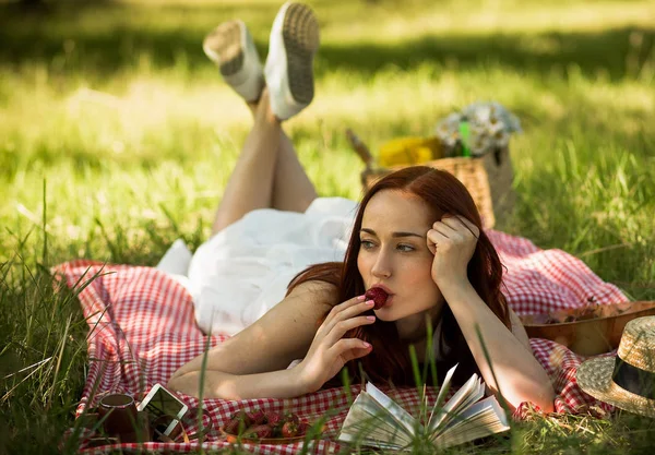 Capelli rossi signora sdraiata sul velo su erba mangiare fragola e leggere un libro . — Foto Stock