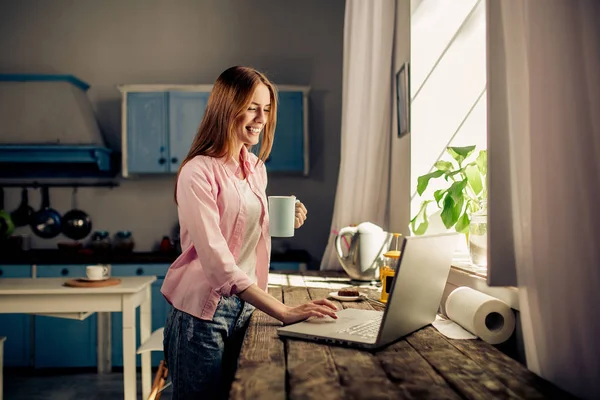Ritratto di bella ragazza in piedi in cucina tastiera sul computer portatile, in possesso di tazza . — Foto Stock