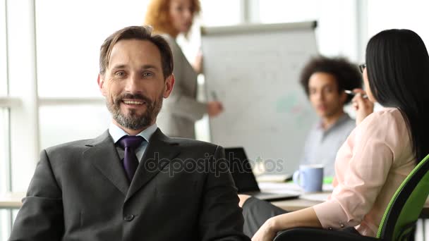 Hombre de negocios sonriente sentado en la oficina con la espalda a sus compañeros de trabajo . — Vídeos de Stock