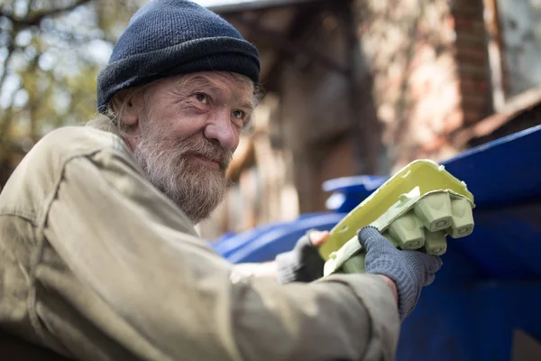 Dirty homeless man holding packing for eggs, standing by the trash can. — Stock Photo, Image