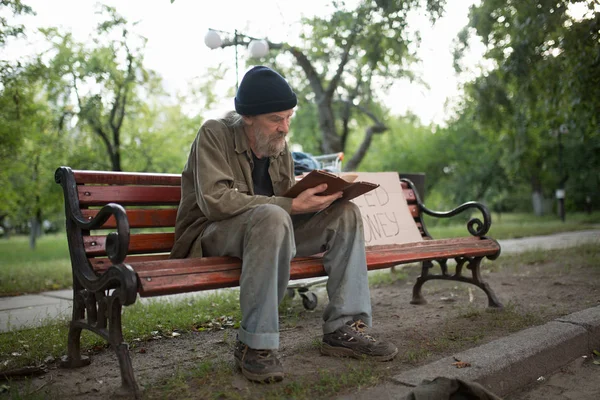 Бездомний чоловік сидить на лавці тримає книгу в руках . — стокове фото