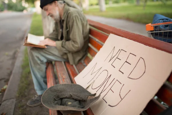 On front view board with sign need money and hat with some coins, reading tramp on backdrop. — Stock Photo, Image