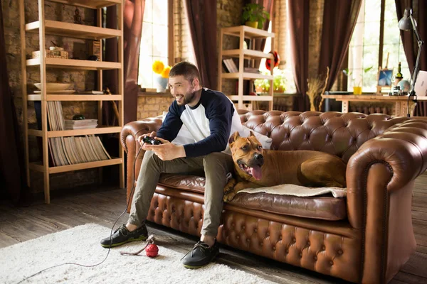 Chico guapo sentado en el sofá con su perro, jugando videojuego . — Foto de Stock