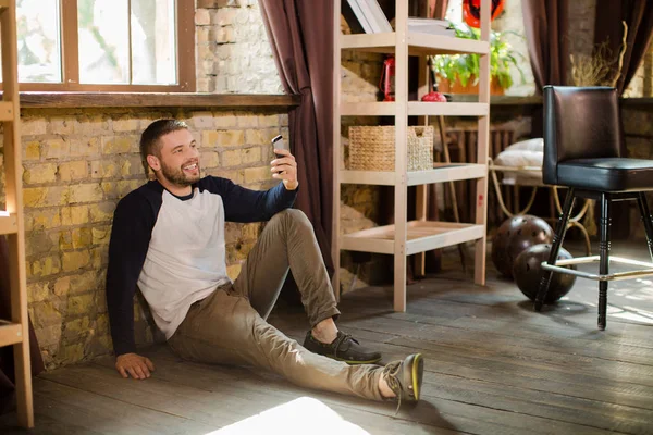 Hombre sonriente en ropa casual en el suelo en casa, sosteniendo el teléfono celular en la mano . — Foto de Stock