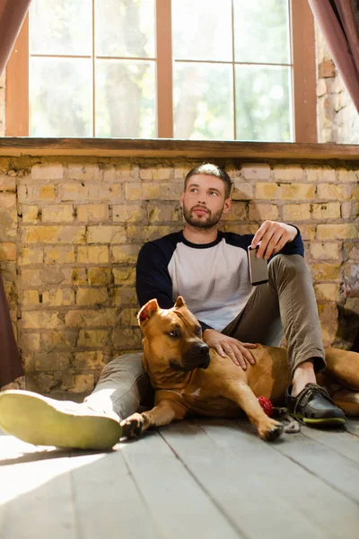 Nadenkende man zit op de vloer met zijn hond, telefoon in de hand houden. — Stockfoto