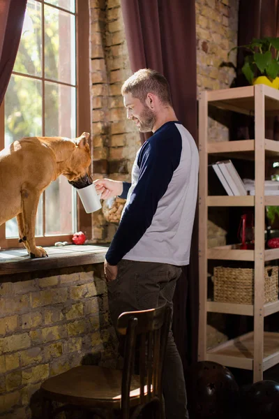 Brutal hombre bebiendo café junto a la ventana, su perro mirando en su taza . — Foto de Stock