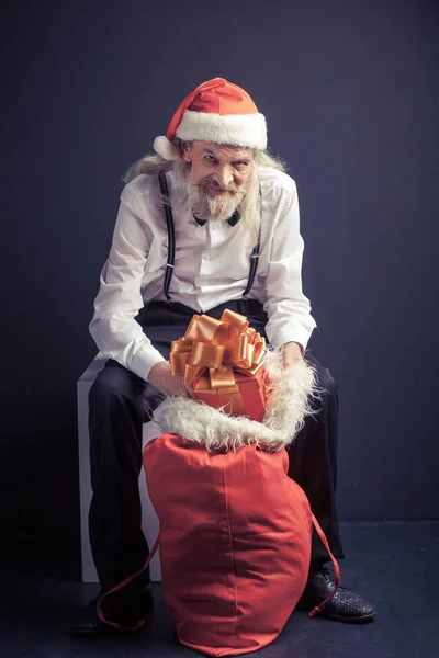 Gubbe i Santa hatt sitter på stol med gåva väska. — Stockfoto