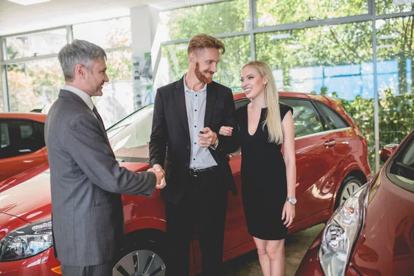 Pasangan bahagia di showroom otomatis, berjabat tangan dengan dealer, mengambil kunci dari mobil baru — Stok Foto