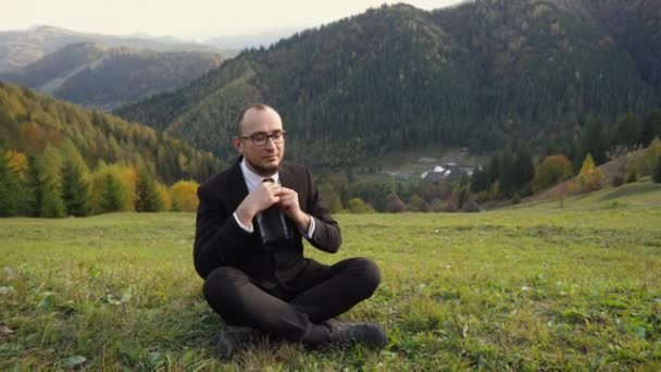 Trabajador de oficina respirando profundamente y meditando mientras está sentado en las montañas con bosque en el fondo — Vídeo de stock