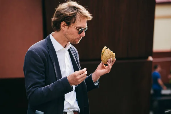 Succesvolle en knappe jongeman in kantoorkleding die een broodje eet op straat tijdens de lunch — Stockfoto