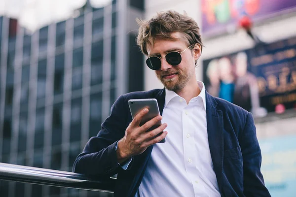 En ung fyr med blondt hår i solbriller ser på sin mobiltelefon, mens han er udendørs på en sommerdag - Stock-foto