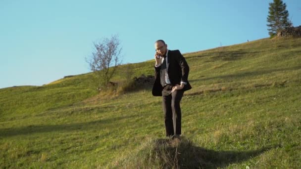 Een man in een zwart pak spreekt op een mobiele telefoon staande op de heuvel — Stockvideo