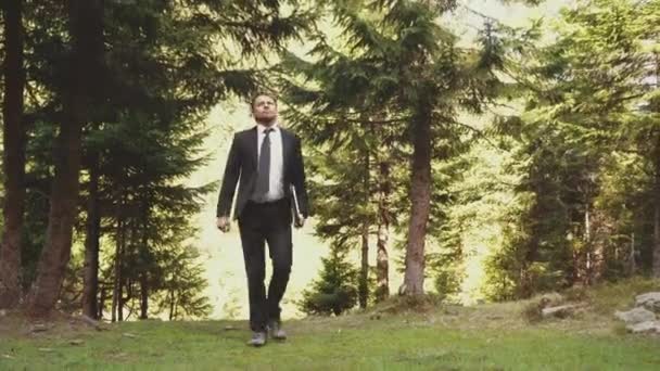 Ein Mann im schwarzen Anzug und weißem Hemd geht durch den Wald, einen Laptop in der Hand. — Stockvideo