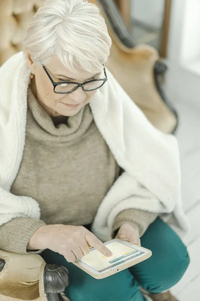 Vieja con gafas está sentada en casa en el sofá de cuero con una tableta en la mano — Foto de Stock