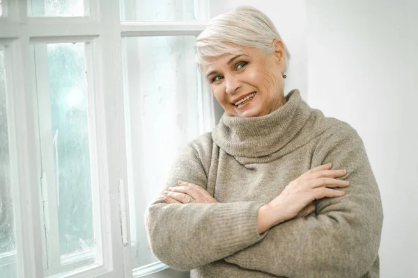 Gris de pelo alto mujer de pie en la ventana y mirando asid —  Fotos de Stock