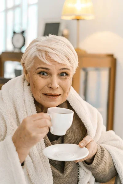 Mulher velha de cabelos grisalhos bebendo chá quente e relaxando em casa — Fotografia de Stock