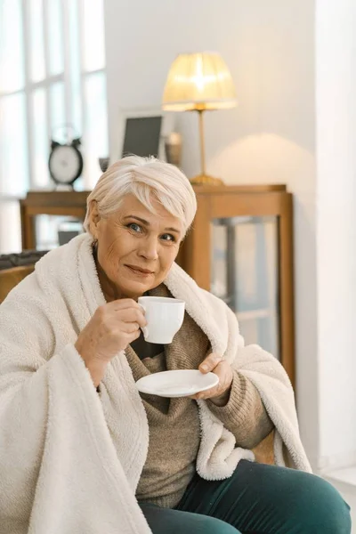 Веселая пожилая женщина, завернутая в теплый вязаный плед, расслабляется дома — стоковое фото