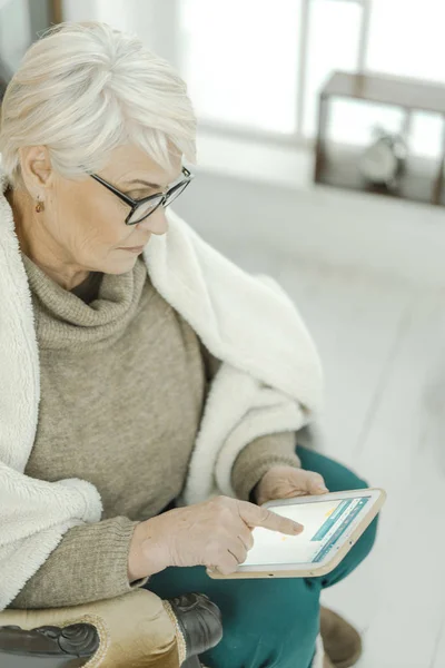 Vieja con gafas está sentada en casa en el sofá de cuero con una tableta en la mano — Foto de Stock