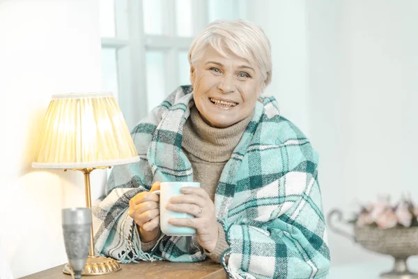 Sorrindo velha senhora embrulhada em uma xadrez xadrez está segurando uma xícara de chá . — Fotografia de Stock