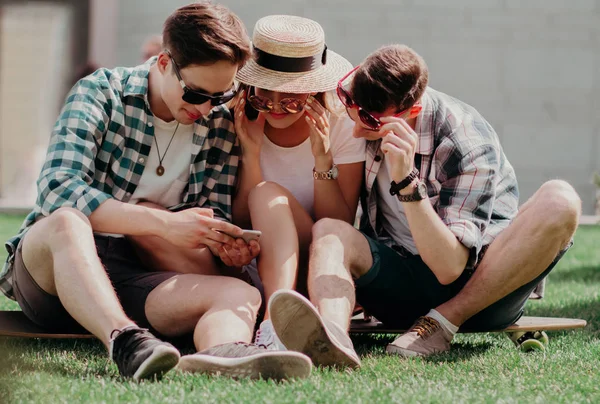 Три молоді друзі сидять на траві на відкритому повітрі і дивляться на мобільний телефон . — стокове фото